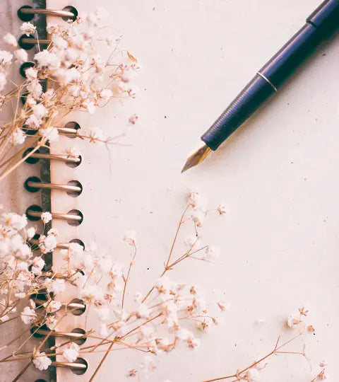 Una pluma fuente negra con punta dorada y flores secas de color blanco rosado esparcidas sobre las páginas abiertas de un cuaderno de espiral con el fondo en blanco.