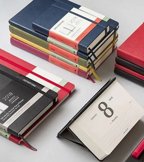  Una vista en ángulo de varios planificadores y cuadernos Moleskine de 2018 apilados y abiertos, mostrando diferentes colores de portada y formatos de calendario.