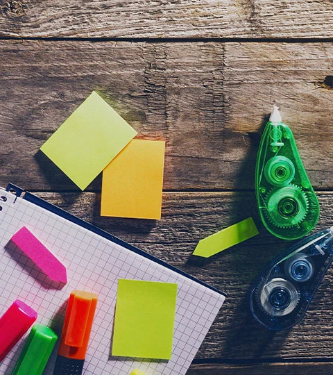 Una vista cenital de un escritorio de madera con un cuaderno de cuadrícula, varios post-its de colores, marcadores fluorescentes y dos dispensadores de cinta correctora.